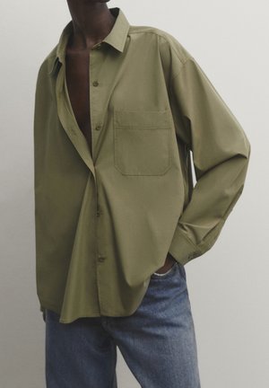 Person wearing oversized olive green button-up shirt with chest pocket and blue jeans, hand in pocket, against plain light background.