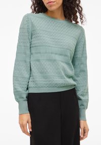 Vrouw draagt een lichtgroene gebreide trui met diamant- en streeppatronen, gecombineerd met zwart, hoog getailleerde broek, staand tegen een witte achtergrond.