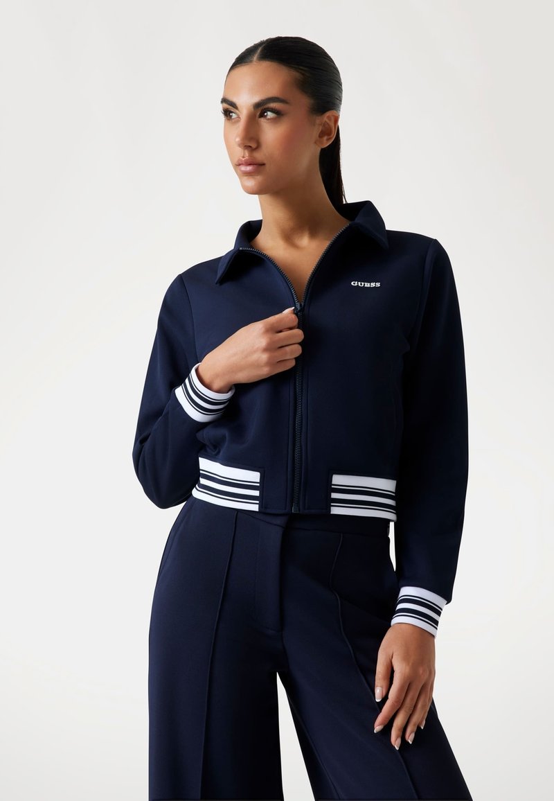 Marineblauwe cropped jacket met ritssluiting aan de voorkant, contrasterende witte gestreepte manchetten en tailleband. Glad stof met een klein logo op de borst.