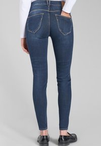 Jeans ajustados de denim azul oscuro con costuras visibles, dos bolsillos traseros y un diseño ceñido, combinados con mocasines negros.