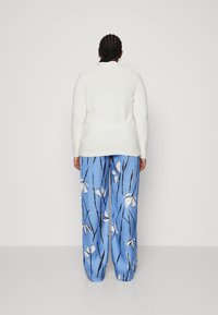Persona con el cabello trenzado que lleva puesta una chaqueta de punto blanca y pantalones de pierna ancha azules con patrones florales blancos y líneas negras, de pie con la espalda mirando hacia afuera.