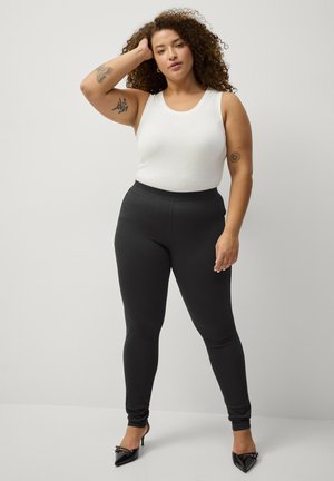 Witte mouwloze top gecombineerd met strakke zwarte leggings, met een gladde textuur en hoge tailleband, aangevuld met puntige zwarte hakken.