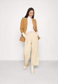 Beige wide-leg trousers paired with a fitted white turtleneck and a brown blazer. The look is completed with beige ankle boots.