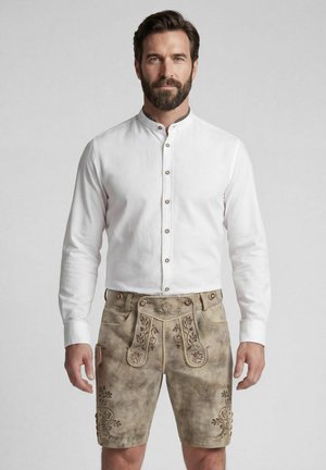 Mann mit dunklem Haar und Bart, der ein weißes Hemd mit Knöpfen und traditionelle beige Lederhosen mit Blumenstickerei trägt.