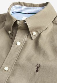 Chemise en coton marron avec un col boutonné, des boutons en nacre et un petit logo de cerf brodé sur la poitrine. Col intérieur rayé.