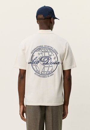 Beige poloshirt med korte ærmer, navy globusgrafik og tekst på ryggen, med en afslappet pasform og ribstrikket krave.