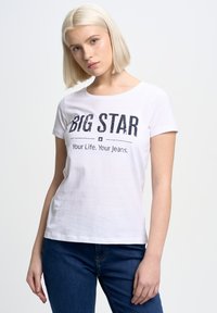 Bílý bavlněný tričko s potiskem "BIG STAR" v tmavém písmu. Krátké rukávy, kulatý výstřih a volný střih. V kombinaci s modrými džínovými kalhotami.