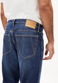 Jean en denim avec un lavage bleu foncé, présentant des poches arrière distinctes, des accents brodés et une étiquette en cuir beige avec du texte embossé.