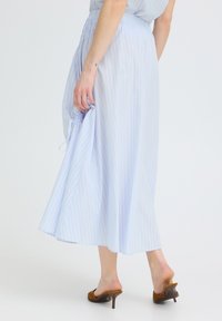Femme portant une robe midi rayée bleu clair, tenant le côté de la jupe, debout pieds nus dans des mules marron à talons sur fond blanc.