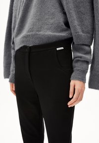 Šedý pletený svetr s širokými rukávy, doplněný černými kalhotami s knoflíkovým zapínáním a bočními kapsami. Štítek viditelný na kalhotách.