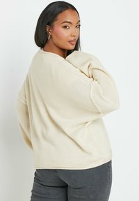 Beige stickad tröja med avslappnad passform, neddragna axlar och strukturerad tyg. Kombinerad med mörkgrå jeans.