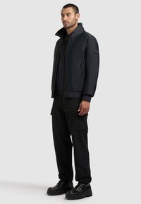Chaqueta bomber negra con cuello alto, cierre de cremallera y bolsillos laterales; combinada con pantalones cargo negros y botas negras gruesas.