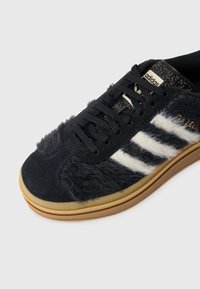 Shoe atlético preto com textura de camurça e pelo, apresenta riscas brancas tradicionais, atacadores pretos e sola de borracha. Logótipo na língua.