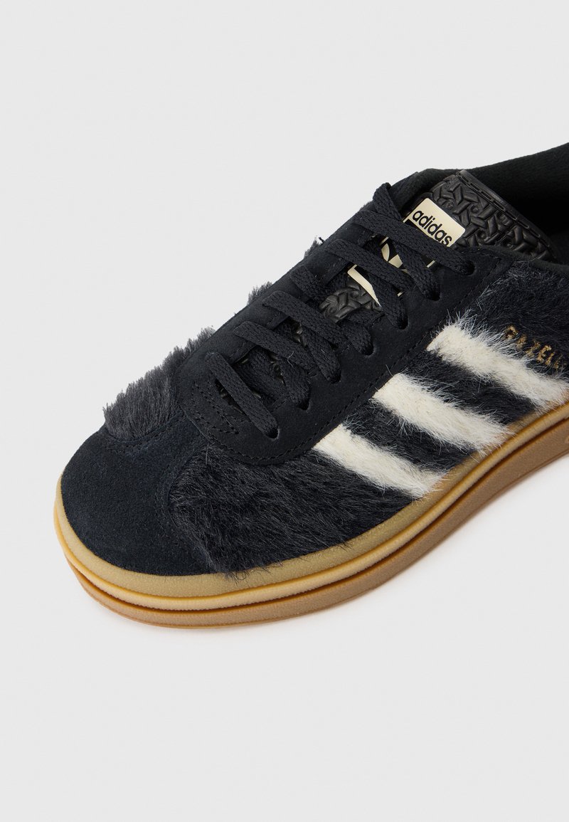 Schwarzer Sportschuh mit Wildleder- und Felltextur, ausgestattet mit traditionellen weißen Streifen, schwarzen Schnürsenkeln und Gummisohle. Logo auf der Zunge.