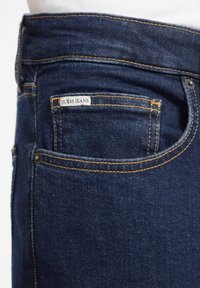 Jeans oscuros con costuras naranjas, que presentan un bolsillo trasero con una etiqueta de "Guess Jeans". Textura de denim suave, diseño clásico.