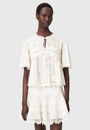 Femme portant un chemisier en dentelle blanche à manches courtes avec lacets et une jupe en dentelle assortie sur un fond uni.