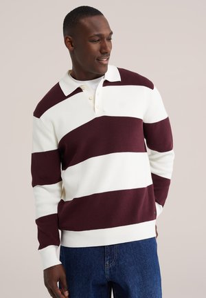 Homme portant un polo à manches longues à larges rayures blanches et rouge foncé, et un jean bleu, regardant sur le côté avec une main dans la poche.
