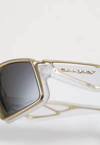Nærbillede af solbriller med guldramme og gradient mørke linser samt Oakley-logo på klare arme mod en hvid baggrund.