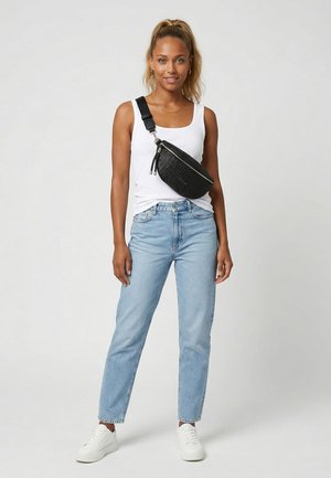 Frau in weißem Tanktop, hellblauen Jeans, weißen Sneakers und einer schwarzen Umhängetasche steht vor einem schlichten grauen Hintergrund.