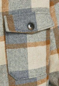 Camicia in flanella a scacchi grigia, crema e ruggine con taschino frontale, caratterizzata da un bottone nero e un motivo di tessuto testurizzato.