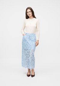 Γυναίκα όρθια με το ένα χέρι στην τσέπη, φορώντας ένα κρεμ μπλουζάκι με μακριά μανίκια και φούστα midi από γαλάζια δαντέλα με λουλούδια, με σκίσιμο μπροστά.