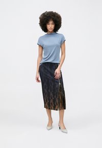 Donna con capelli ricci che indossa una maglia blu con maniche a cappuccio, una gonna midi nera a gradiente con stampa di serpente e cerniera laterale, e tacchi grigi a punta.