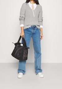 Sac fourre-tout en nylon noir avec des poignées courtes, présentant une forme structurée et un design minimaliste. Porté avec un jean bleu clair et un pull gris.