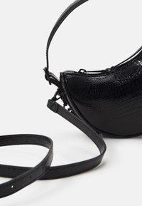 Sac à main noir avec une finition texturée en motif crocodile. Présente une forme arrondie, une longue bandoulière réglable et des détails de design minimalistes.