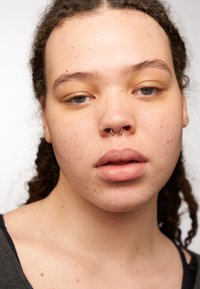 Närbild av ett ansikte med lockigt hår, septumpiercing, ljus hy med fräknar och neutral smink som framhäver de naturliga dragen.