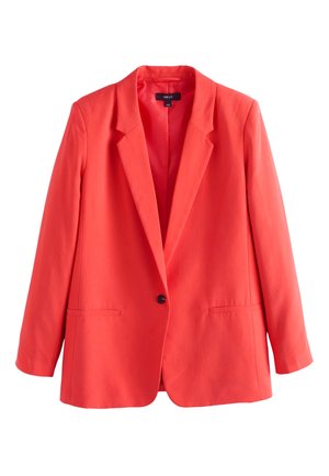 Ung kvinde med krøllet hår iført en orange dragt over en hvid polkaprikket top med blonder, stående udendørs med hænderne i lommerne.
