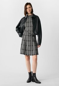 Giacca oversize in pelle nera abbinata a un vestito a motivi a scacchi bianco e nero. La modella indossa stivaletti neri alla caviglia. Sfondo semplice.