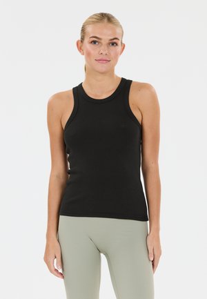 Kvinde med blondt hår i en hestehale iført en sort ærmeløs ribbet tanktop og lysegrå leggings, stående mod en ensfarvet hvid baggrund.