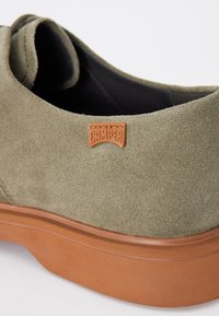 Pantof suede verde măsliniu cu o talpă din cauciuc bej. Prezintă un emblemă din piele maro pe lateral. Design modern și minimalist.