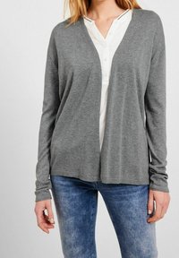 Grå långärmad kofta med V-ringning, tillverkad av mjukt stickat material. Har ribbade ärmslut och nederkant. Bärs över en vit skjorta med knappar.