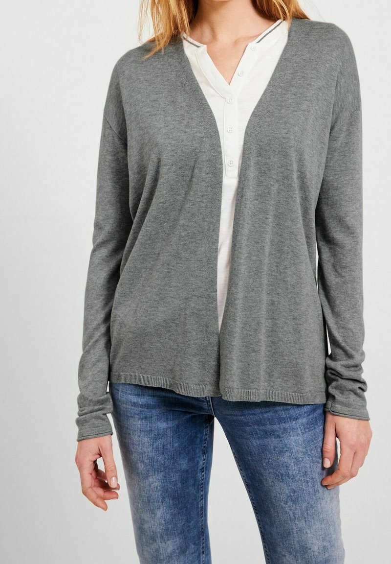 Grå långärmad kofta med V-ringning, tillverkad av mjukt stickat material. Har ribbade ärmslut och nederkant. Bärs över en vit skjorta med knappar.