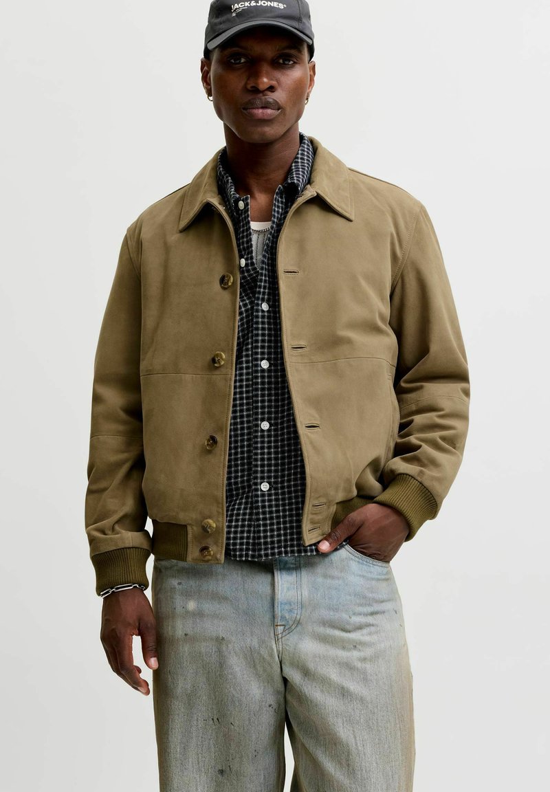 Homme portant une veste boutonnée vert olive, une chemise à carreaux noir et blanc, un jean bleu clair délavé et une casquette noire avec un logo blanc.