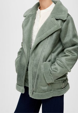 Vrouw in een groene schapenvachtjack met ruime revers, ritssluiting aan de voorkant, gespsdetails en een wit shirt eronder, poseert met handen in de zakken.