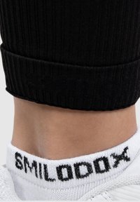 White sock featuring black "SMILODON" lettering at the cuff, paired with a black ribbed fabric ankle sleeve. Textured surface visible.