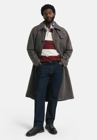 Grauer Hahnentrittmantel mit großem Kragen, darunter ein kastanienbraun-weiß gestreiftes Polohemd, blaue Jeans und schwarze Schnürschuhe. Stehende Pose.