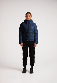 Blå puffy jakke med høj krave, to lynlåslommer foran og et logo på venstre bryst. Sammen med sorte joggers og sko.