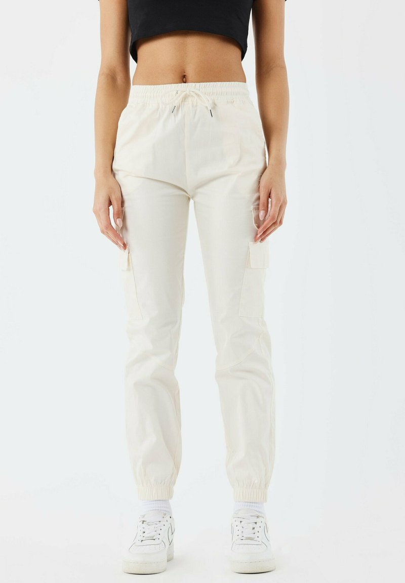 Cremfarbene Cargo-Hosen mit einem elastischen Bund und Bündchen, ausgestattet mit Seitentaschen und einer glatten Stoffstruktur. Kombiniert mit weißen Sneakers.