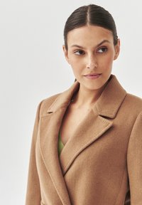 Abrigo de lana color camello con cuello chal, que presenta un diseño de un solo botón y una textura suave, superpuesto a una blusa verde.