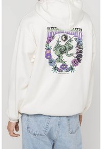 Bílá mikina s kapucí se zelenou žábou na přední straně obklopenou barevnými květinovými vzory a designem oka na zadní straně, vyrobená z měkkého materiálu.