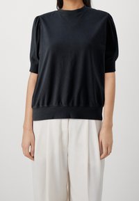 Suéter negro con mangas cortas abullonadas y cuello redondo. Hecho de una tela suave, presenta un dobladillo y puños acanalados. Combinado con pantalones claros.