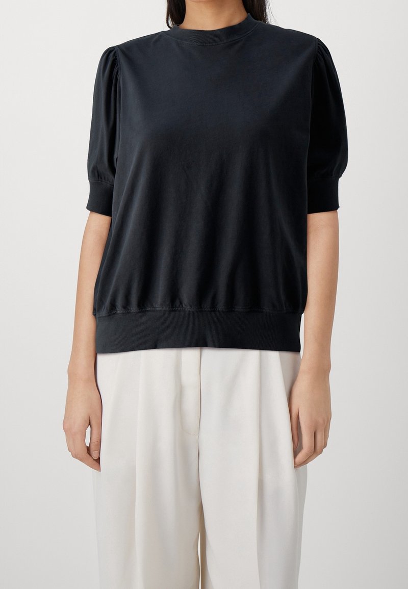 Suéter negro con mangas cortas abullonadas y cuello redondo. Hecho de una tela suave, presenta un dobladillo y puños acanalados. Combinado con pantalones claros.
