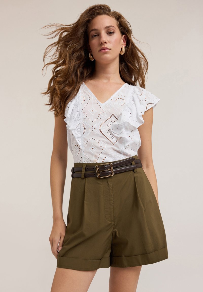 Weiße, geraffte Bluse mit Lochmuster, kombiniert mit olivgrünen, knielangen Shorts und einem braunen Gürtel mit rechteckiger Schnalle.