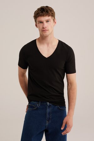 Mladić s kratkom kovrčavom kosom, obučen u crnu majicu s V-izrezom i plave traperice, stoji ispred jednobojne svijetle pozadine.