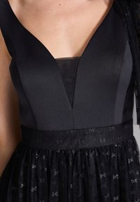 Robe noire avec un décolleté en V profond, corsage structuré et jupe texturée présentant des motifs subtils. Ceinture fine en matériau assorti.