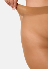 Pantys transparentes en un color nude, con una textura suave y fina, que cuentan con una cintura flexible siendo tirada por una mano. No presentan patrones notables ni componentes metálicos.