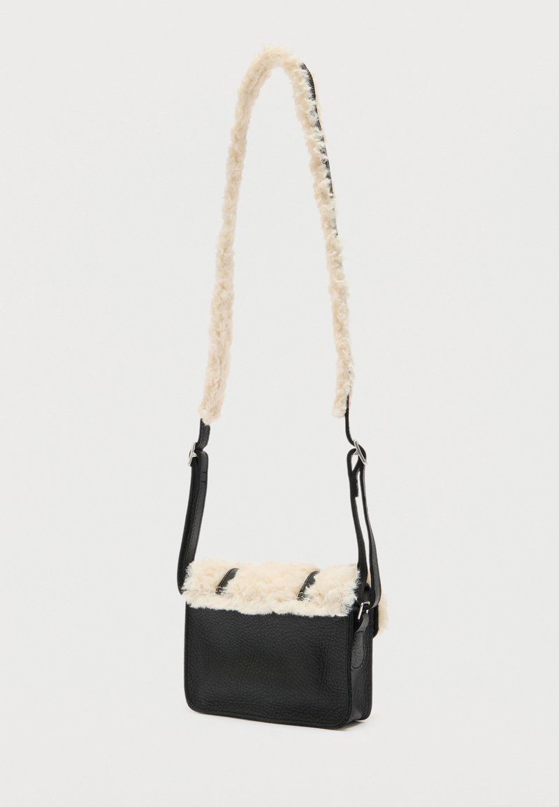 Schwarze Leder-Umhängetasche mit cremefarbenem, weichem Fleecebesatz an Klappe und Riemen vor weißem Hintergrund.